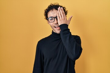 Hispanic man standing over yellow background covering one eye with hand, confident smile on face and surprise emotion.