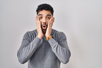 Hispanic man with beard standing over white background afraid and shocked, surprise and amazed expression with hands on face