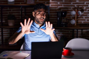Hispanic man with beard using laptop at night afraid and terrified with fear expression stop gesture with hands, shouting in shock. panic concept.