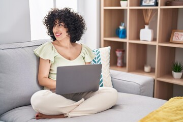 Young brunette woman with curly hair using laptop sitting on the sofa at home looking away to side with smile on face, natural expression. laughing confident.