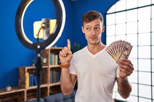 Caucasian man recording vlog tutorial with smartphone at home holding money smiling with an idea or question pointing finger with happy face, number one