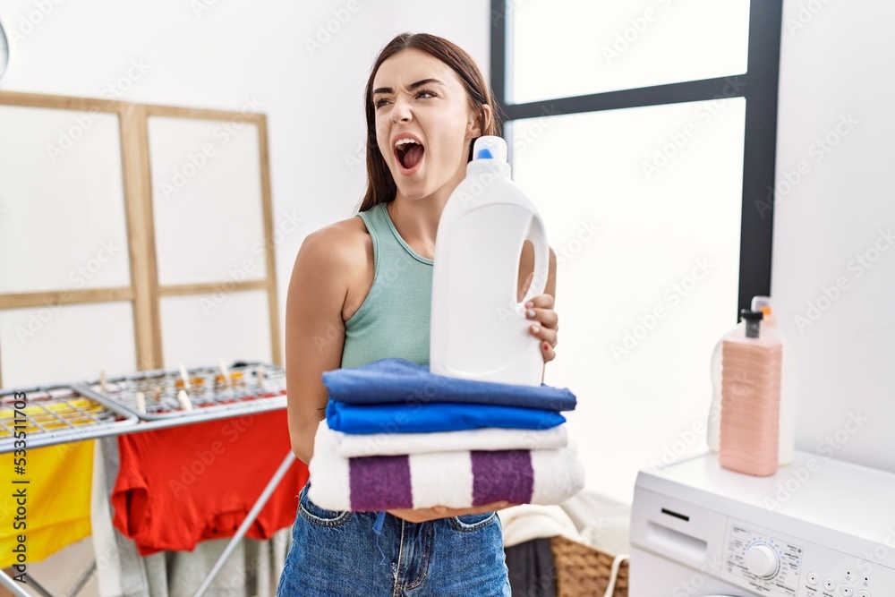 Sticker young hispanic woman doing laundry holding detergent bottle and folded clothes angry and mad screami