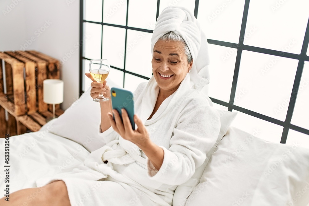 Poster Middle age grey-haired woman drinking champagne and using smartphone at bedroom.