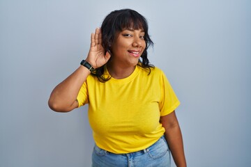 Hispanic woman standing over blue background smiling with hand over ear listening an hearing to rumor or gossip. deafness concept.