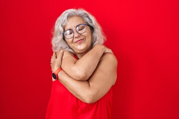 Middle age woman with grey hair standing over red background hugging oneself happy and positive, smiling confident. self love and self care