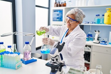 Middle age woman wearing scientist uniform pouring liquid on test tube at laboratory