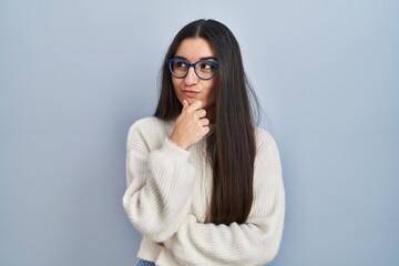 Young hispanic woman wearing casual sweater over blue background looking confident at the camera with smile with crossed arms and hand raised on chin. thinking positive.