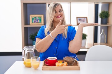 Obraz na płótnie Canvas Caucasian plus size woman eating breakfast at home amazed and smiling to the camera while presenting with hand and pointing with finger.