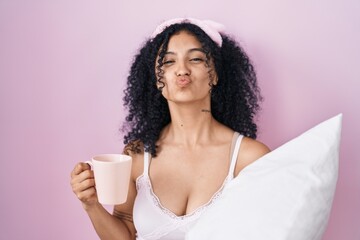 Hispanic woman with curly hair wearing pajama drinking a cup of coffee looking at the camera blowing a kiss being lovely and sexy. love expression.