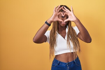 African american woman with braided hair standing over yellow background doing heart shape with hand and fingers smiling looking through sign