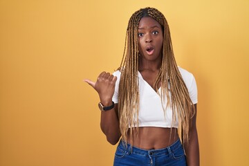 African american woman with braided hair standing over yellow background surprised pointing with hand finger to the side, open mouth amazed expression.