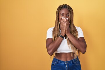 African american woman with braided hair standing over yellow background laughing and embarrassed...