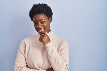 African american woman standing over blue background looking confident at the camera smiling with crossed arms and hand raised on chin. thinking positive.