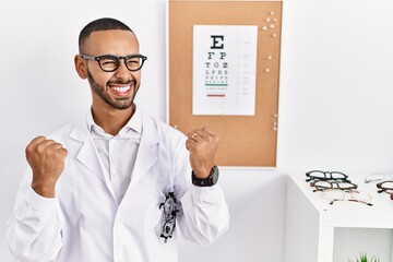 Fototapeta na wymiar African american optician man standing by eyesight test celebrating surprised and amazed for success with arms raised and eyes closed. winner concept.