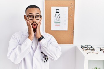 African american optician man standing by eyesight test afraid and shocked, surprise and amazed expression with hands on face