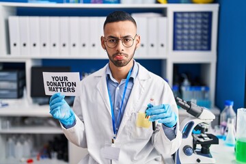 Young hispanic man working at scientist laboratory holding your donation matters holding blood sample depressed and worry for distress, crying angry and afraid. sad expression.