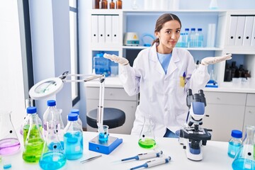 Fototapeta premium Young brunette woman working at scientist laboratory clueless and confused expression with arms and hands raised. doubt concept.
