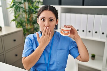 Young latin dentist woman holding denture with braces covering mouth with hand, shocked and afraid...