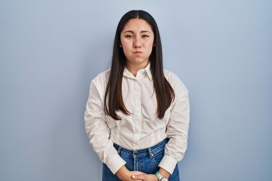 Young latin woman standing over blue background puffing cheeks with funny face. mouth inflated with air, crazy expression.