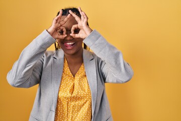 Fototapeta premium African american woman with braids standing over yellow background doing ok gesture like binoculars sticking tongue out, eyes looking through fingers. crazy expression.