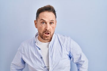 Fototapeta premium Middle age caucasian man standing over blue background in shock face, looking skeptical and sarcastic, surprised with open mouth