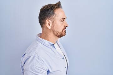 Middle age caucasian man standing over blue background looking to side, relax profile pose with natural face and confident smile.