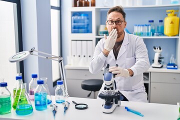 Middle age caucasian man working at scientist laboratory covering mouth with hand, shocked and...