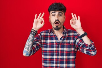 Young hispanic man with beard standing over red background looking surprised and shocked doing ok approval symbol with fingers. crazy expression - obrazy, fototapety, plakaty