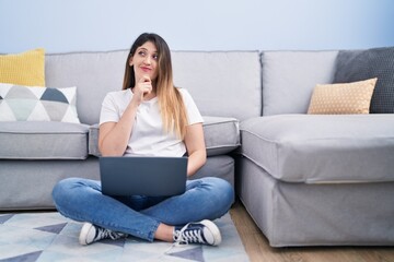 Obraz premium Young brunette woman sitting on the floor at home using laptop serious face thinking about question with hand on chin, thoughtful about confusing idea