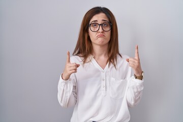 Brunette woman standing over white isolated background pointing up looking sad and upset, indicating direction with fingers, unhappy and depressed.