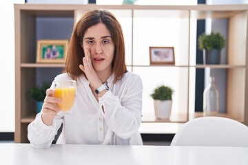 Brunette woman drinking glass of orange juice hand on mouth telling secret rumor, whispering malicious talk conversation
