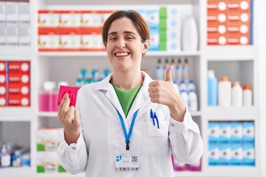Brunette woman working at pharmacy drugstore holding condom smiling happy and positive, thumb up doing excellent and approval sign
