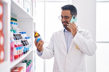 Young hispanic man pharmacist holding medication bottle talking on smartphone at pharmacy