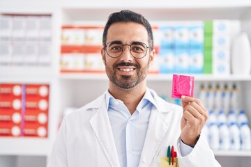 Hispanic man with beard working at pharmacy drugstore holding condom looking positive and happy...