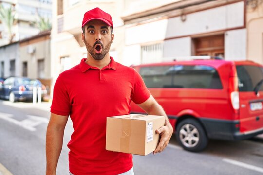 Young hispanic man with beard wearing delivery uniform and cap holding box scared and amazed with open mouth for surprise, disbelief face