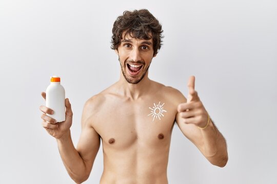Young hispanic man standing shirtless holding sunscreen lotion pointing fingers to camera with happy and funny face. good energy and vibes.