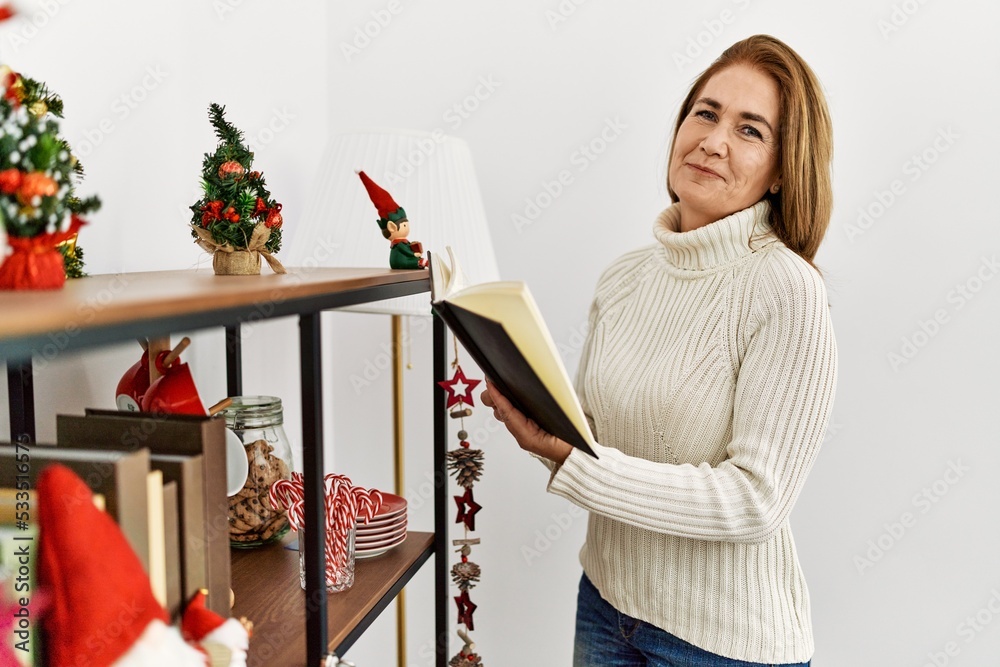 Poster middle age caucasian woman reading book standing by christmas decor at home