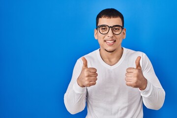 Young arab man wearing casual white shirt and glasses success sign doing positive gesture with hand, thumbs up smiling and happy. cheerful expression and winner gesture.