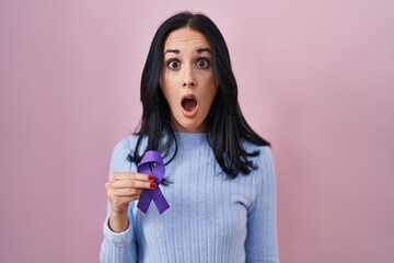 Hispanic woman holding purple ribbon awareness scared and amazed with open mouth for surprise,...