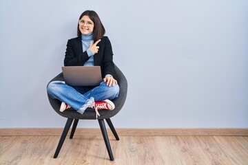 Young hispanic woman sitting on chair using computer laptop cheerful with a smile on face pointing...