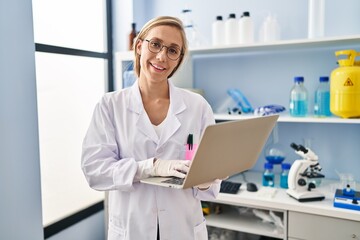 Obraz na płótnie Canvas Young blonde woman scientist smiling confident using laptop at laboratory