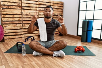 Young indian man sitting on training mat at the gym pointing up looking sad and upset, indicating...