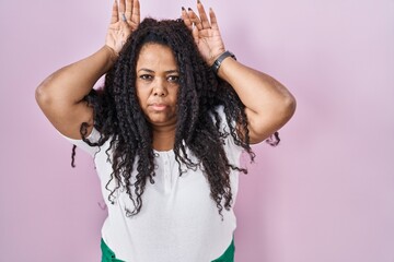 Plus size hispanic woman standing over pink background doing bunny ears gesture with hands palms...