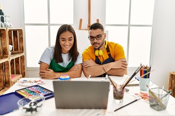 Obraz premium Young hispanic couple using laptop sitting on the table at art studio happy face smiling with crossed arms looking at the camera. positive person.