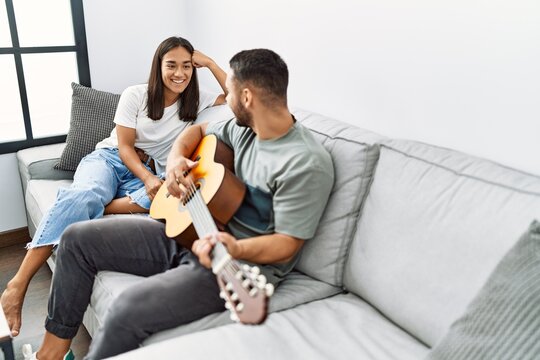 Young Latin Couple Playing Classical Guitar Sitting On The Sofa At Home.