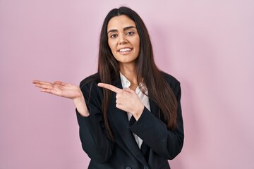 Young brunette woman wearing business style over pink background amazed and smiling to the camera while presenting with hand and pointing with finger.