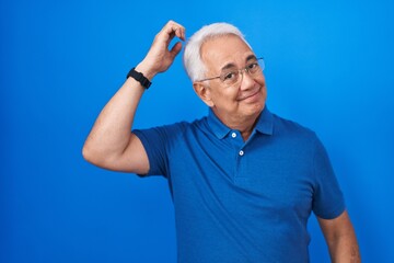 Middle age man with grey hair standing over blue background confuse and wonder about question. uncertain with doubt, thinking with hand on head. pensive concept.