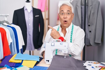 Middle age man with grey hair dressmaker using sewing machine surprised pointing with finger to the side, open mouth amazed expression.