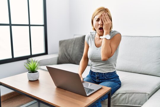 Middle age blonde woman using laptop at home yawning tired covering half face, eye and mouth with hand. face hurts in pain.