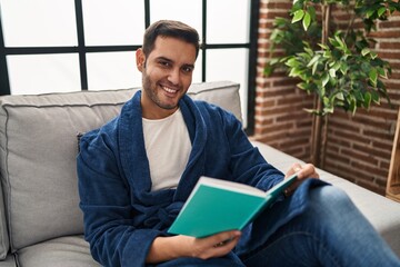 Young hispanic man wearing bathrobe reading book at home